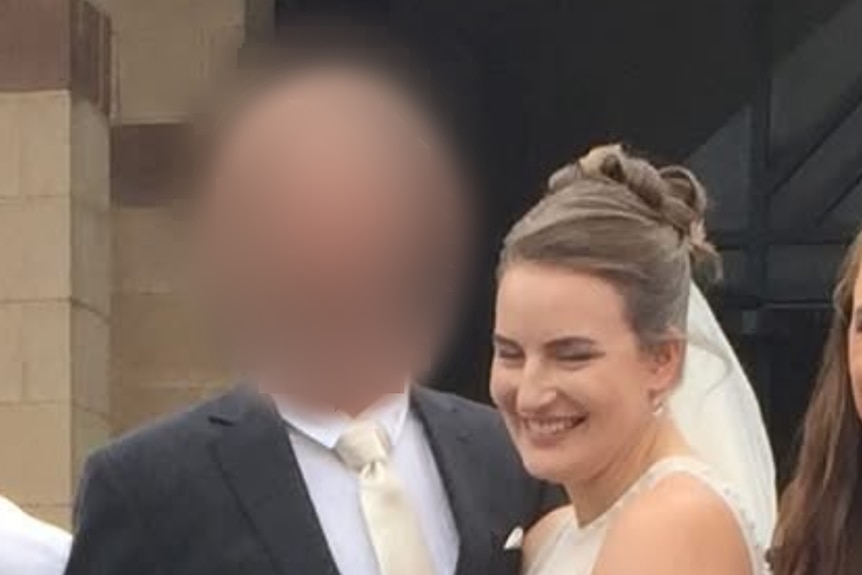 A woman in bridal gown and veil, smiling as she stands next to the groom. His face is blurred
