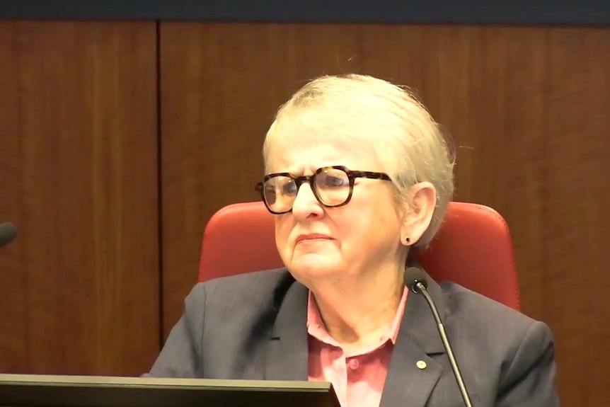 A woman with short blonde hair and brown speckled rimmed glasses in a grey blazer and pink shirt sits behind two microphones.