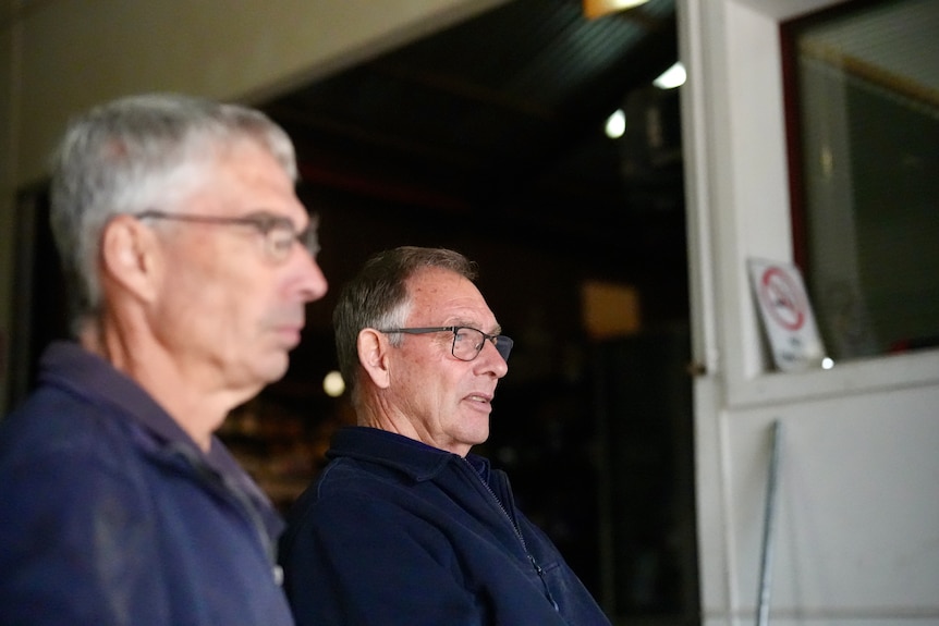 Two middle aged men, one focused one blurred, looking to the right of the camera, inside mechanic workshop