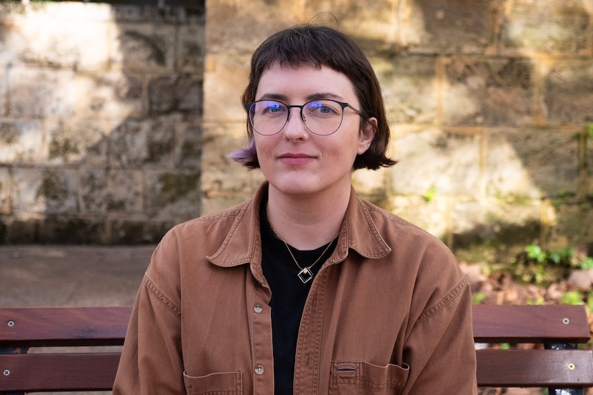 A person with glasses and short hair sits on a bench looking towards the camera.