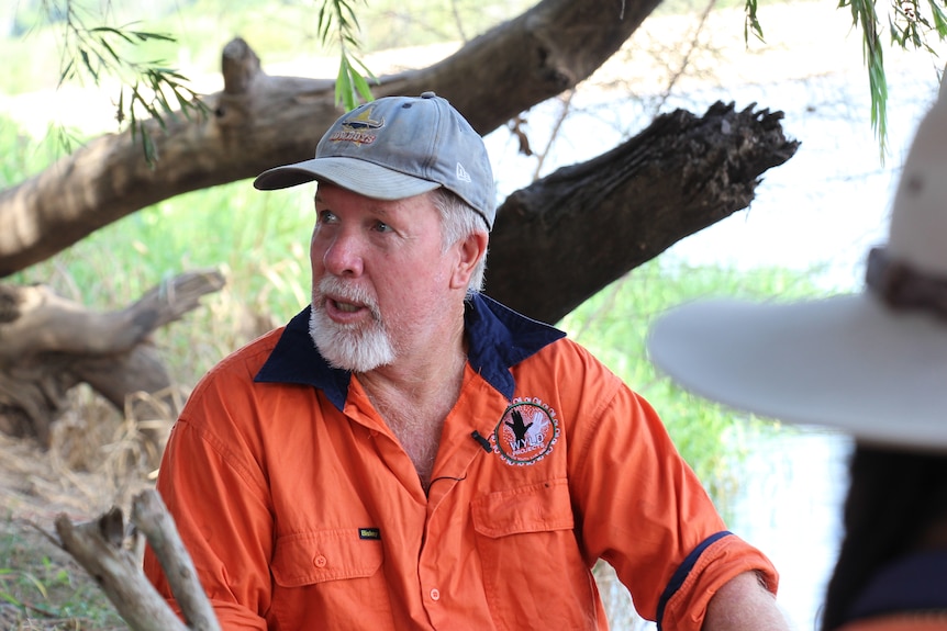 A man with a white beard wearing an orange shirt and hat with a river behind him