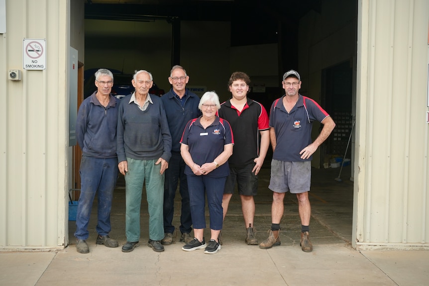 Five men and one woman stand in front of mechanic workshop, smiling