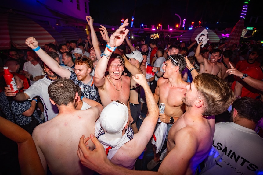 A group of young men with their shirts off jumping and dancing in a bar.