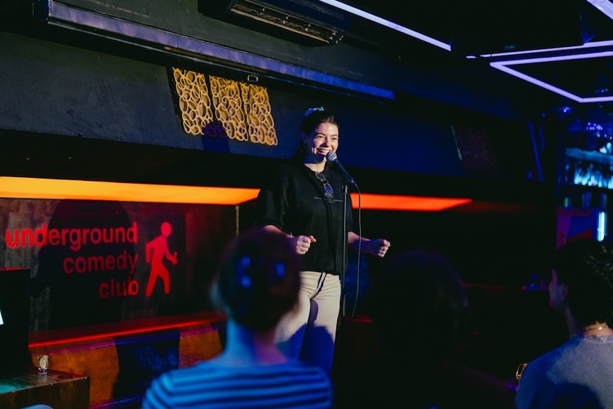 A person stands smiling on a dark stage speaking into a microphone.