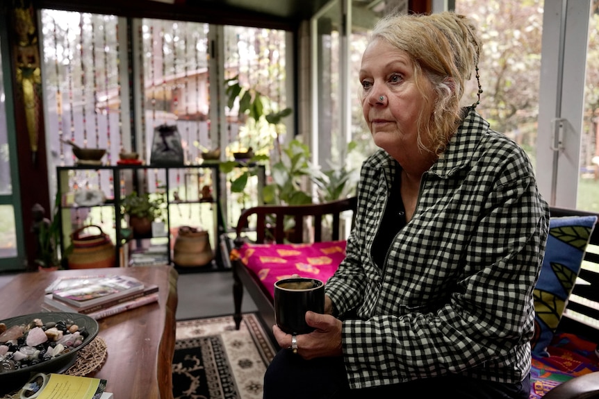 An older woman sits at a couch nursing a cup of tea