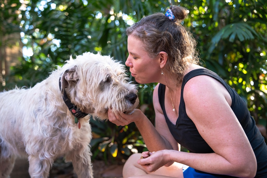 A white woman, blonde / brown hair holding face of her big white dog, pouting to give him a kiss on the head.