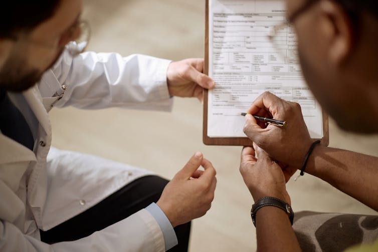 Close up of Black man signing medical form while male doctor guiding paperwork during consultation.