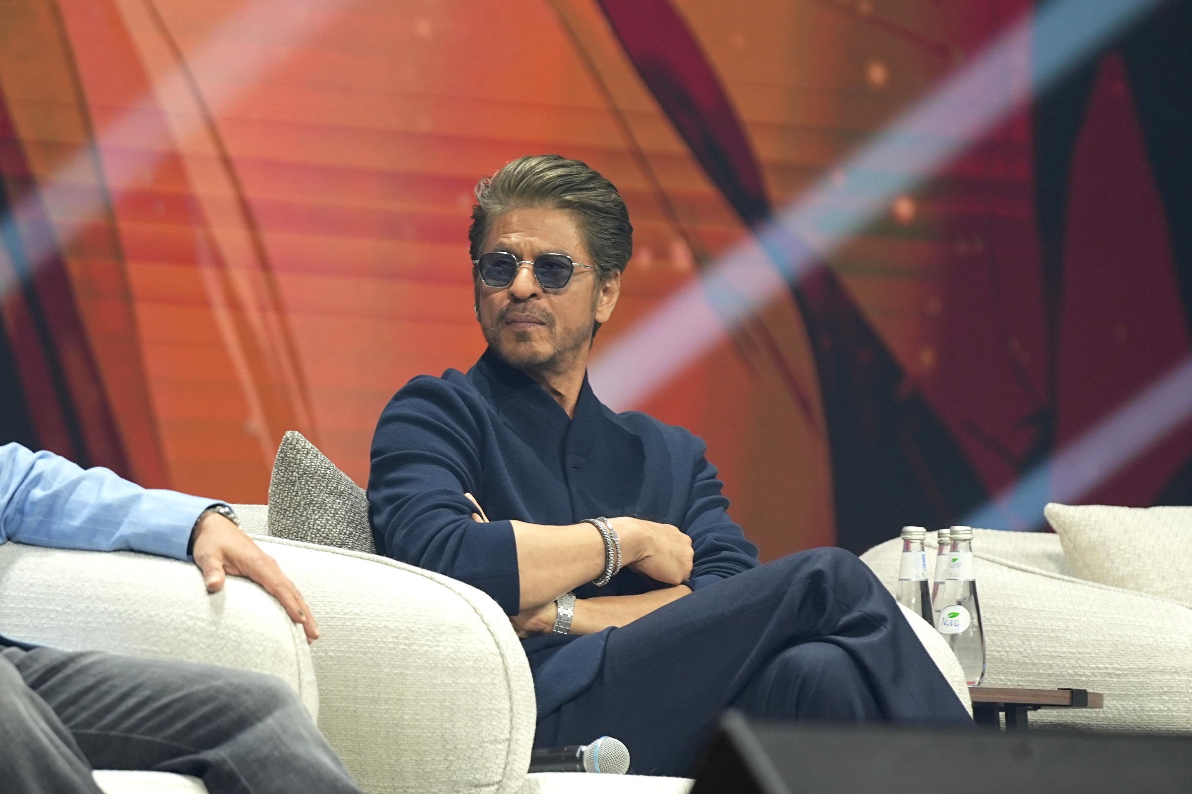 Shah Rukh Khan, in a navy suit and dark glasses, sits in a comfortable chair with an interviewer on stage.