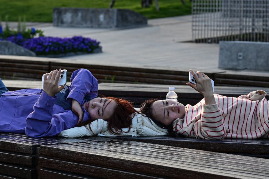 Two women lying on a bench look at their phones along the Huangpu River in Shanghai on April 9, 2026.