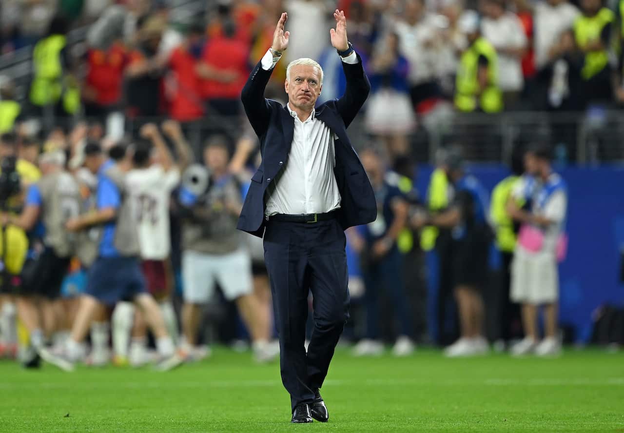 A middle-aged white man in a suit is walking on a football pitch. He has his arms raised as he claps. 