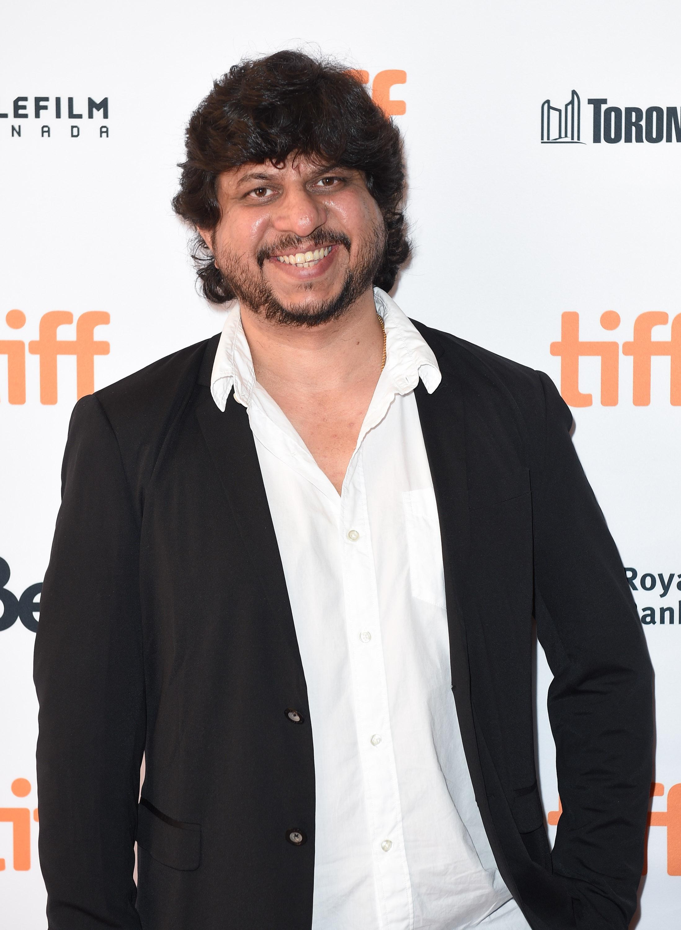 Honey Trehan, a middle-aged man with curly dark hair and beard, in a casual black jacket and open white shirt, in front of a film festival backdrop.