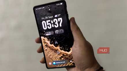 Hand holding a Samsung Galaxy with One UI lock screen, showing date, time, and widgets.