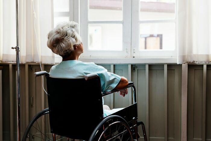 An elderly patient in a wheelchair looks out a hospital window, reflecting hospital horror stories.