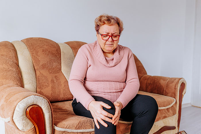 An elderly woman sitting on a sofa, holding her knee in pain, a medical symptom doctors don't want ignored.