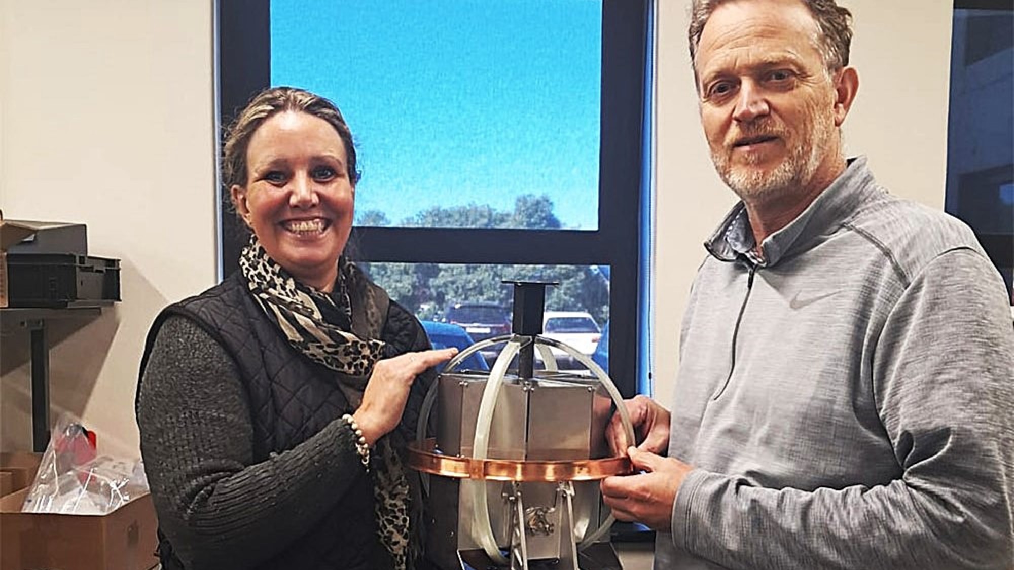 two smiling people pose for a picture next to a spherical metal device
