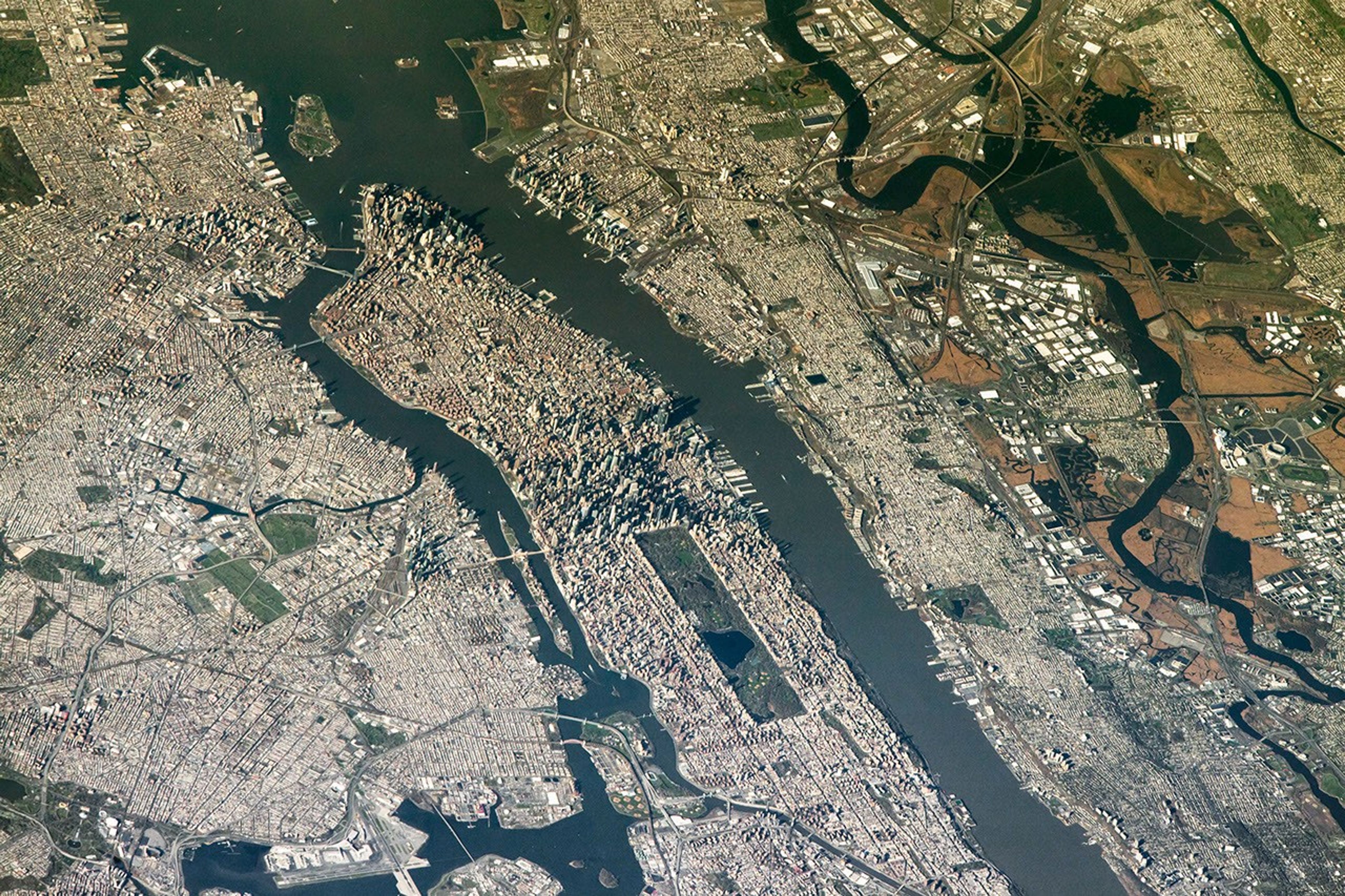An aerial view shows the New York New Jersey Stadium and surrounding Meadowlands on the right, with the dense urban landscape of Manhattan Island centered in the frame.