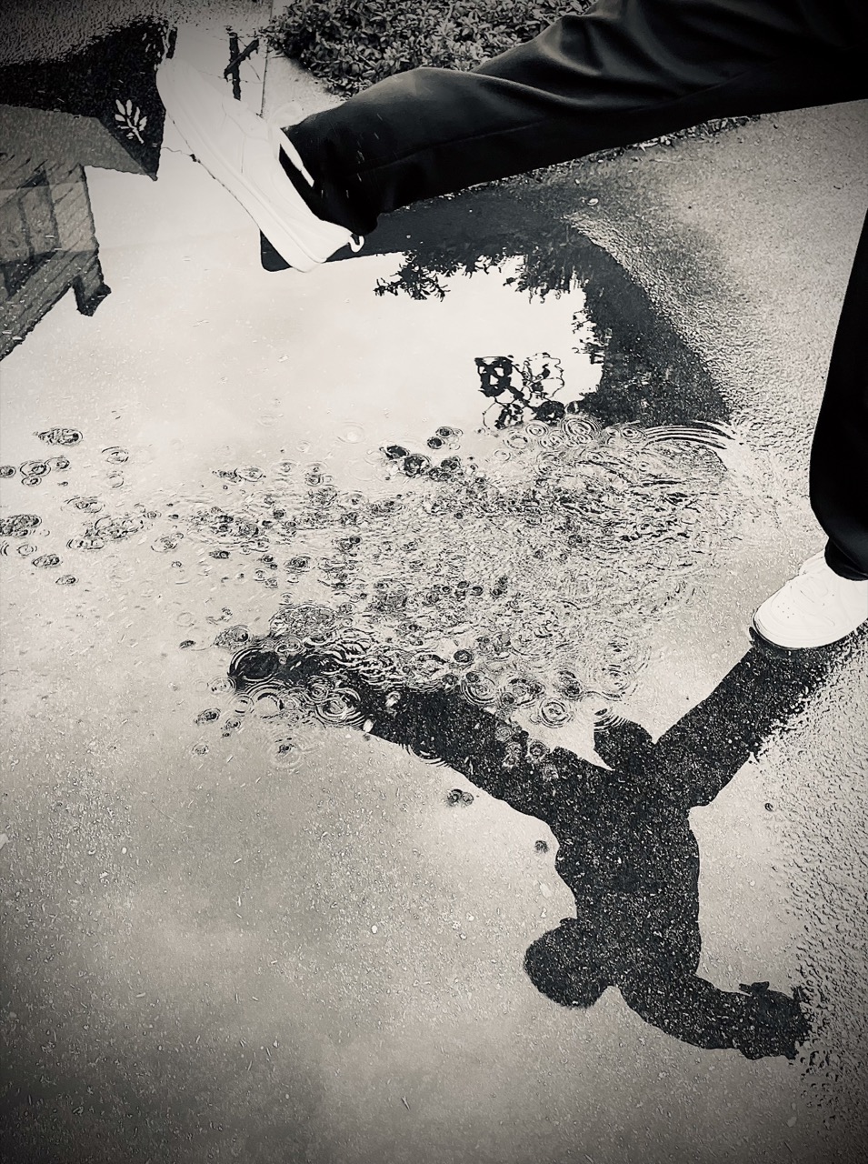 Photograph of a child's reflection in a puddle as they prepare to step over it, image is monochrome