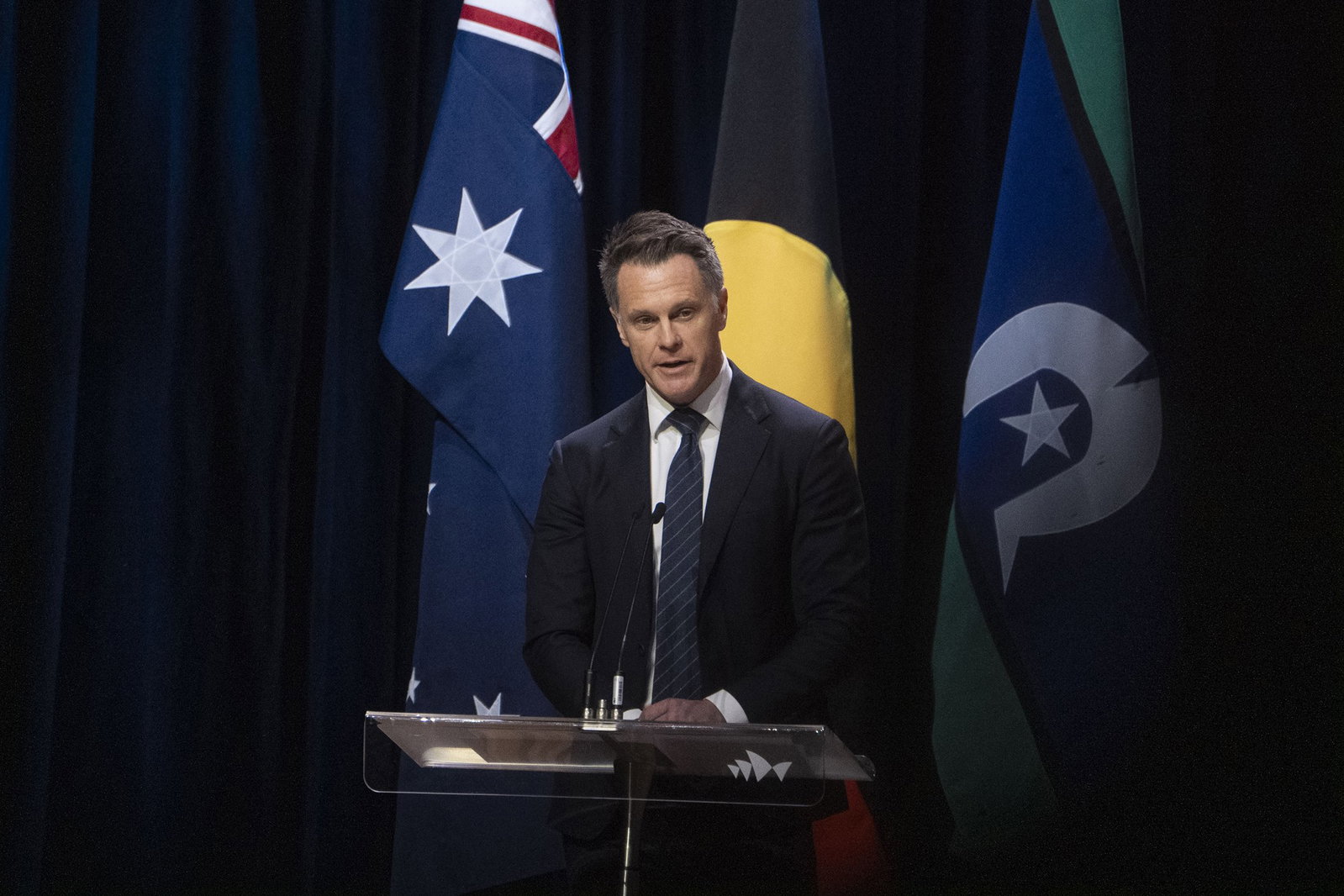 Man in suit talking in front of Australian and Indigenous flags.