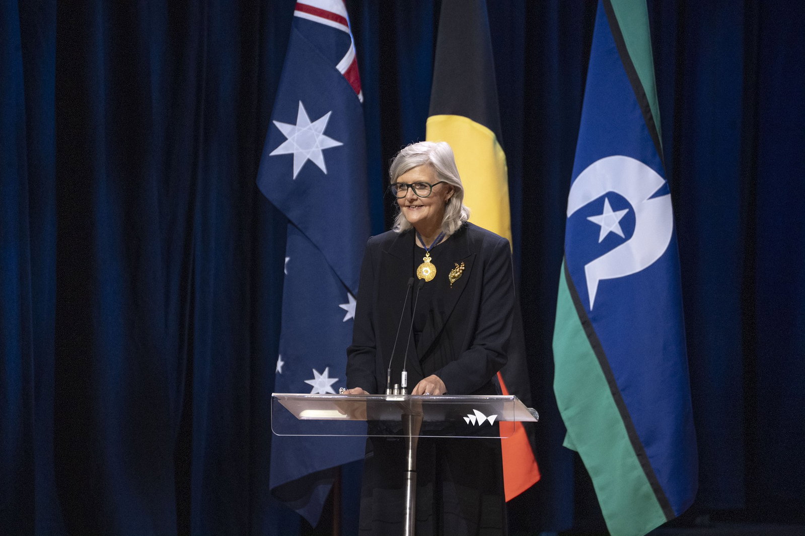 Sam Mostyn with grey hair, black blazer, smiling in front of Australian and Indigenous flags.