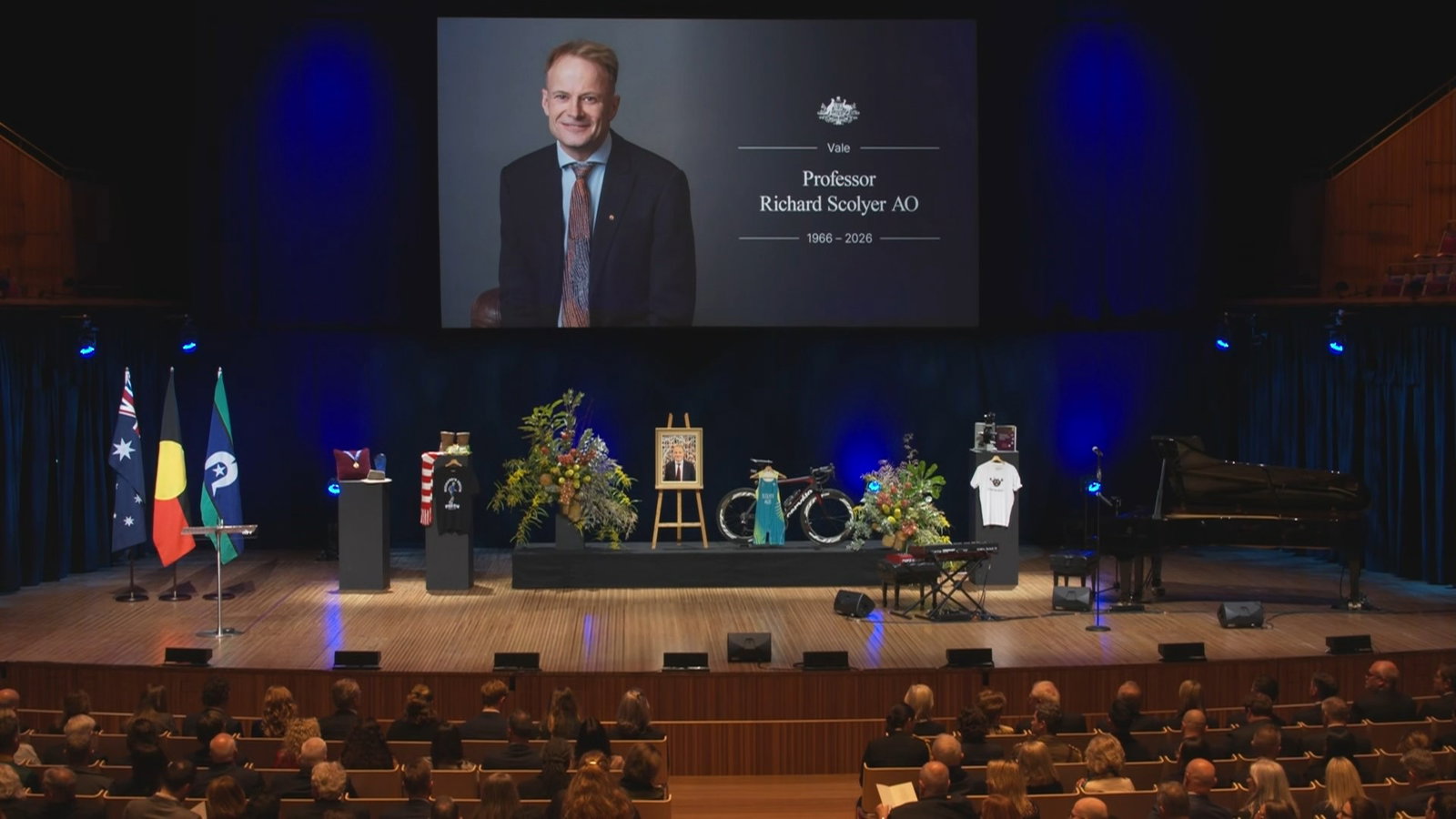 The concert hall with crowd, a stage, and banner of Richard Scolyer.