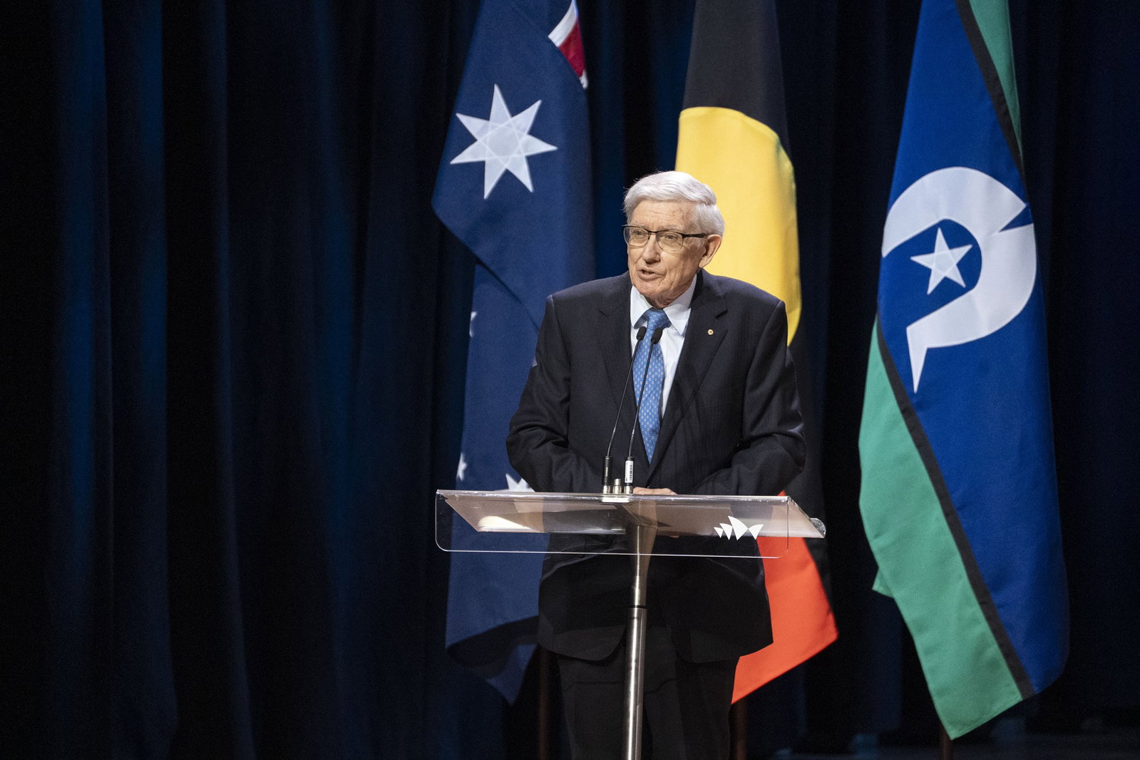 John Thompson in a suit talking in front of an Australian and Indigenous flags