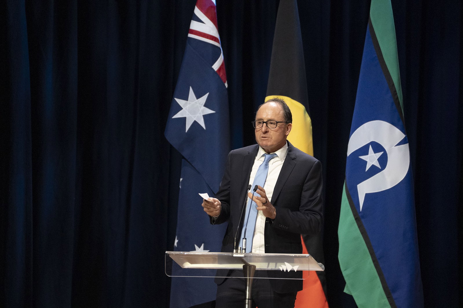 Jim Finlay in a navy suit and blue tie talking in front of Australian and Indigenous flags.