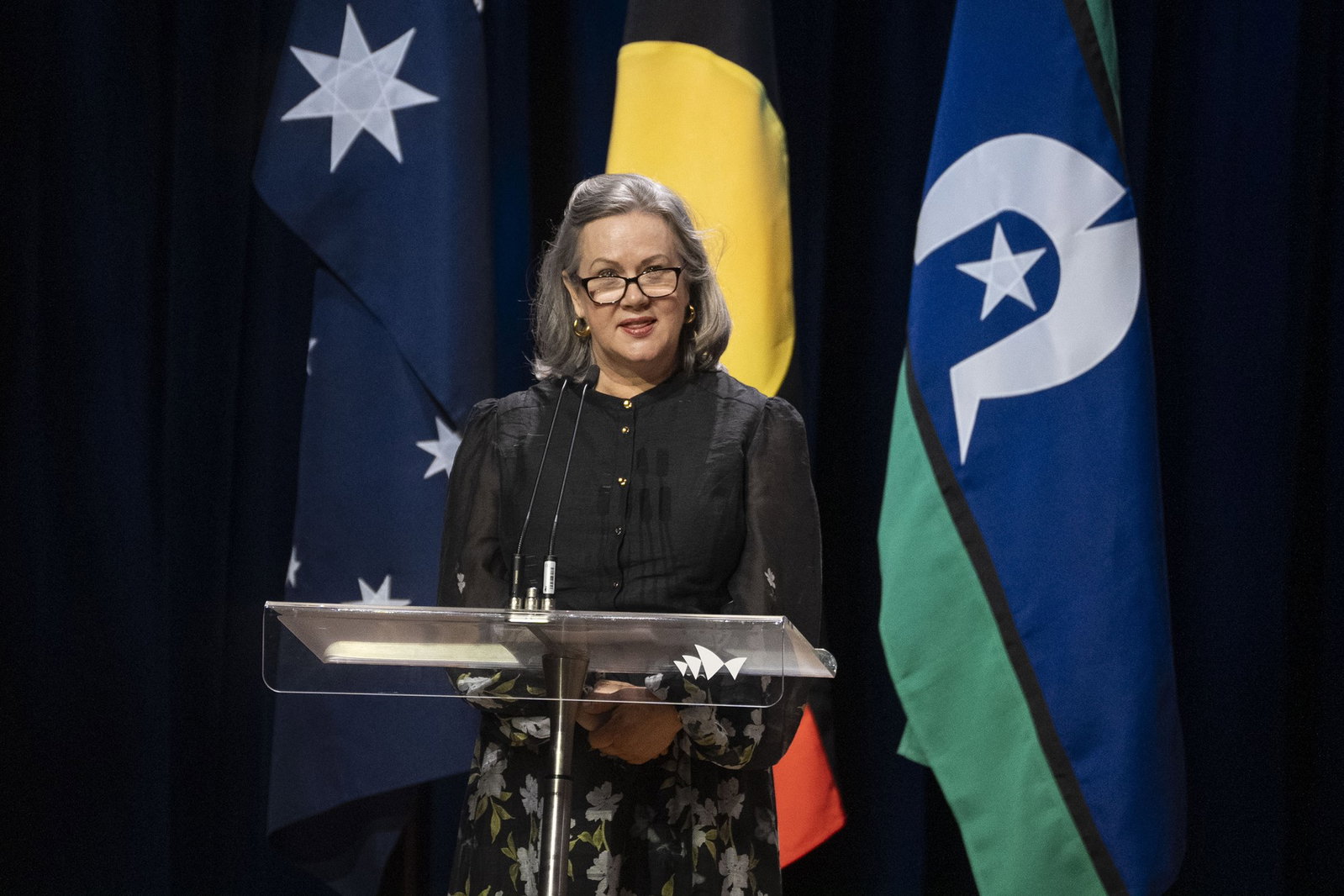 Katie Nicholl in a black dress, speaking in front of Australian and Indigenous flags.