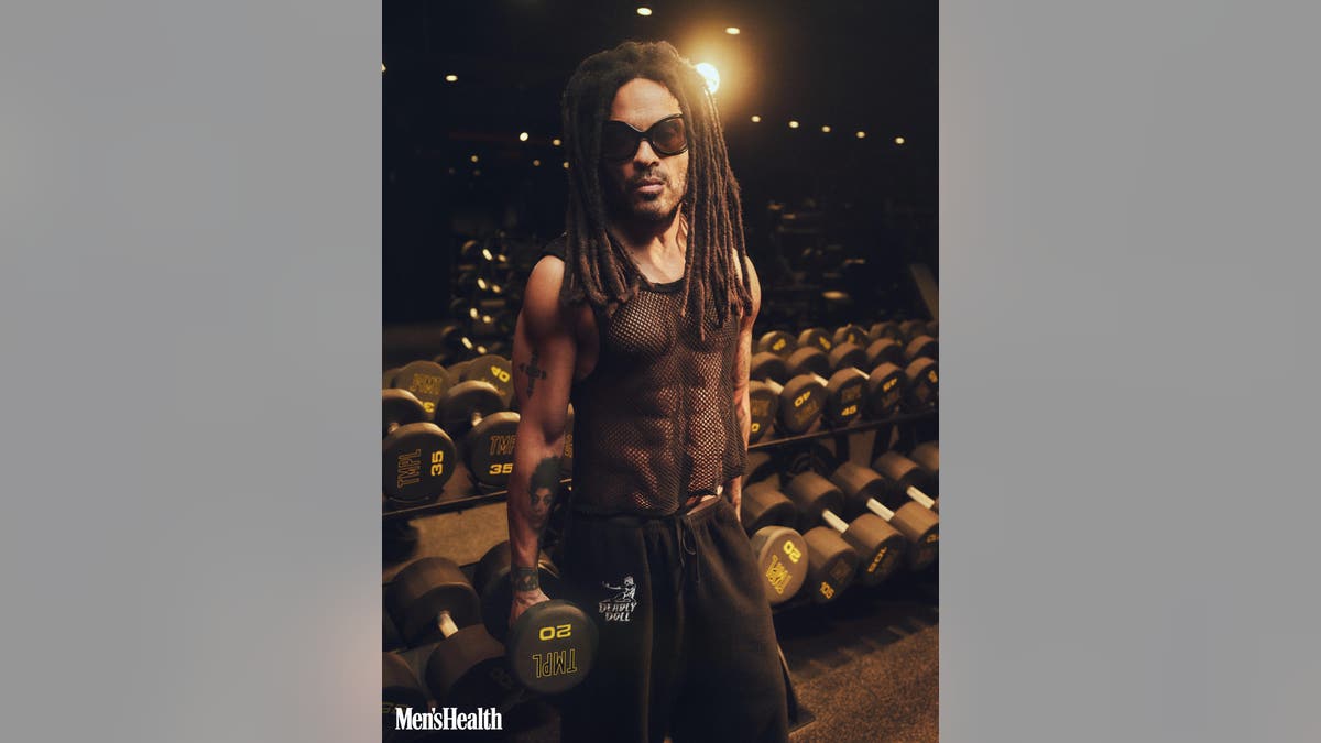 Lenny Kravitz stands holding a dumbbell in a gym with rows of free weights behind him.