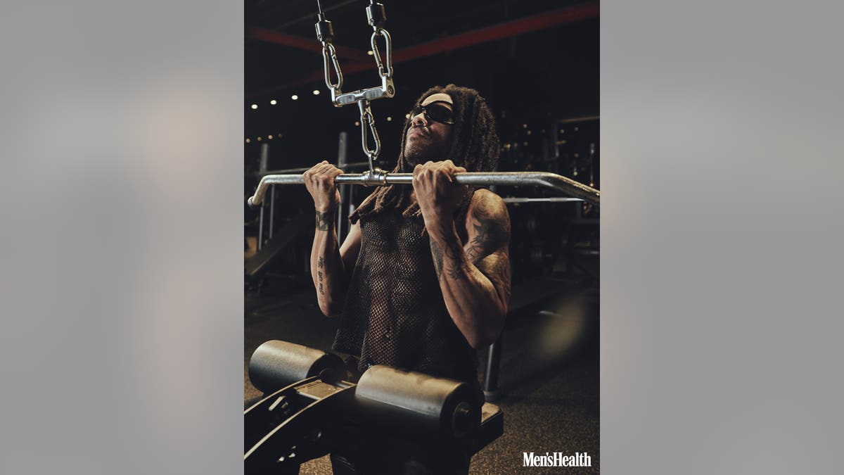 Lenny Kravitz performs a lat pulldown exercise on a weight machine in a gym.