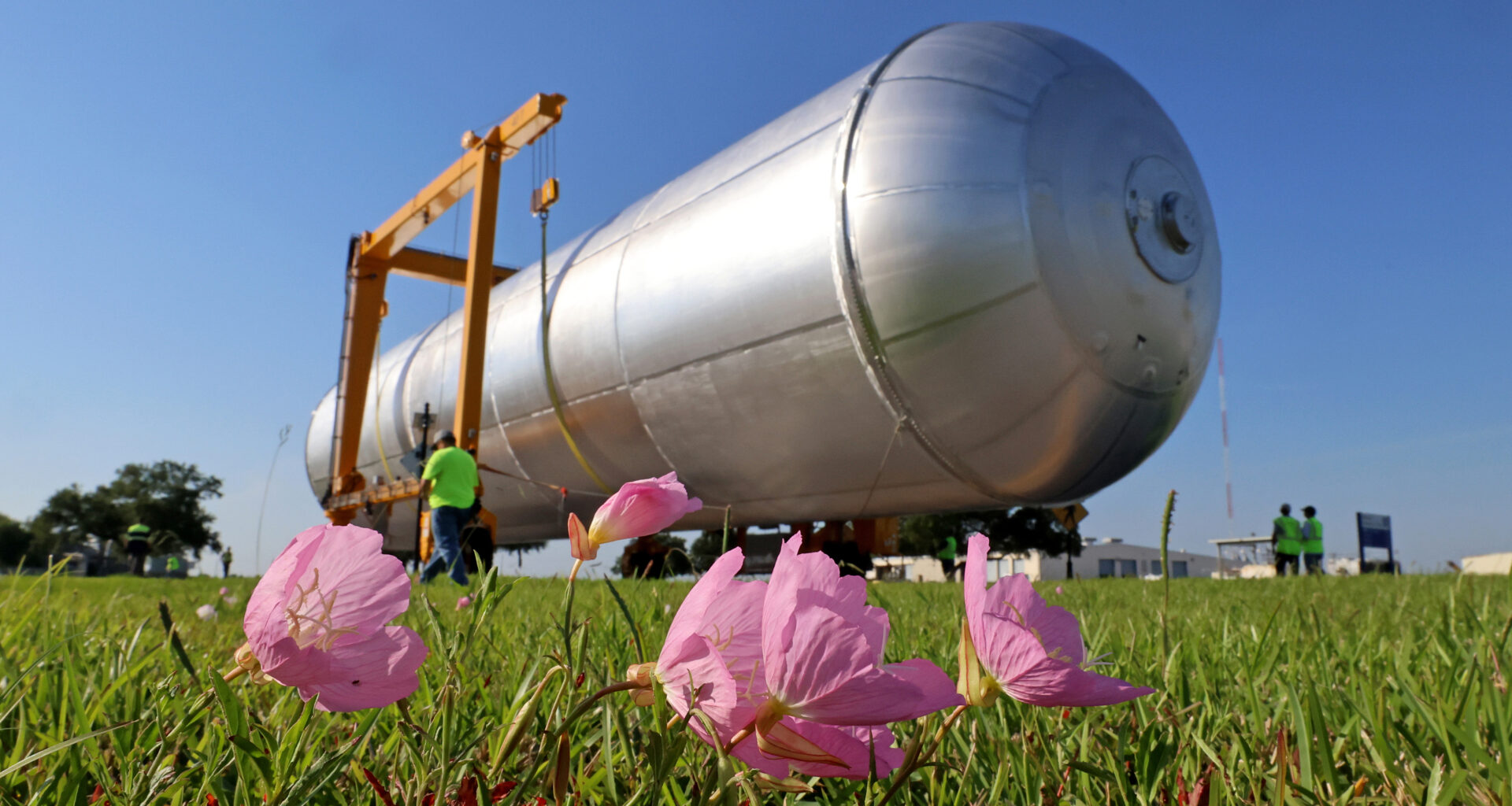 In the foreground, a few pink flowers on a grassy field lean to the right. Behind them is a long, silver cylinder being moved by crews in bright green safety shirts and vests. The massive object is the liquid hydrogen tank for the Artemis IV rocket.