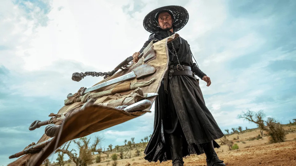 Still from "Blades of the Guardians." Low-angle image of a person in a long black robe and wide-brimmed hat dragging a large bundle of weapons across a barren landscape under a cloudy sky.
