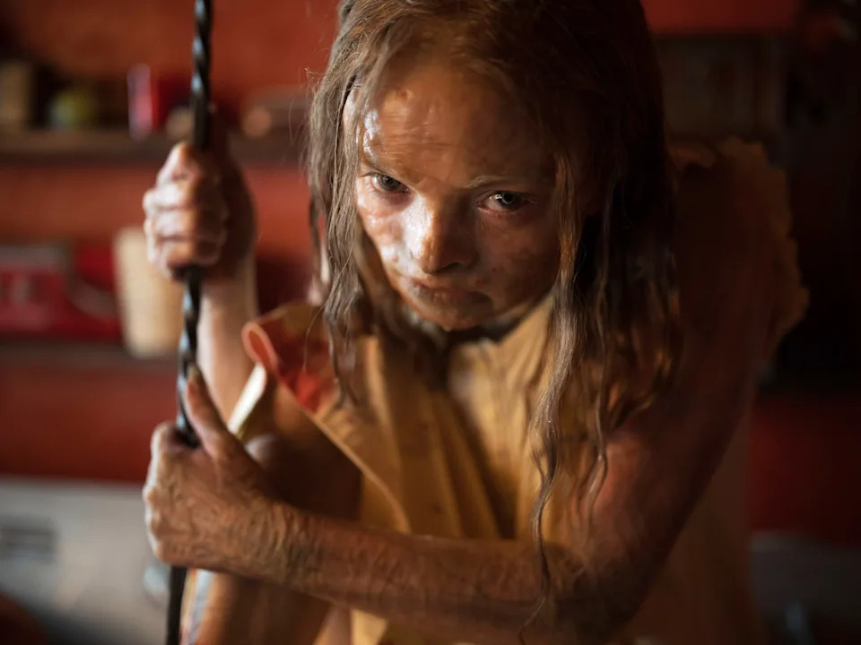 Close-up of a young girl with a dirt-covered face and arms gripping a rope indoors, staring intensely with a serious expression.