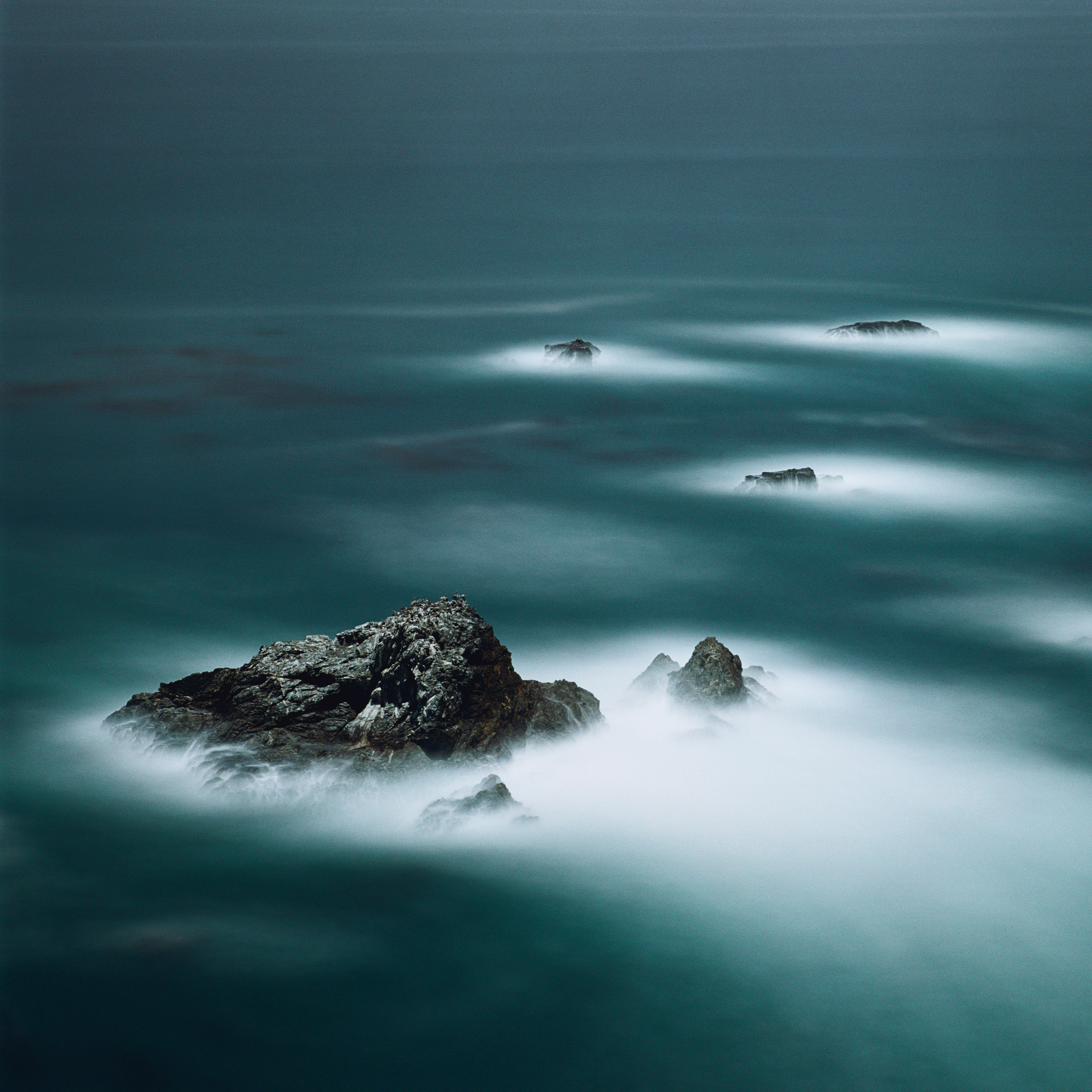 A dramatic seascape capturing dark rocks jutting out of a deep teal ocean, with long-exposure mist swirling around them like white fog.
