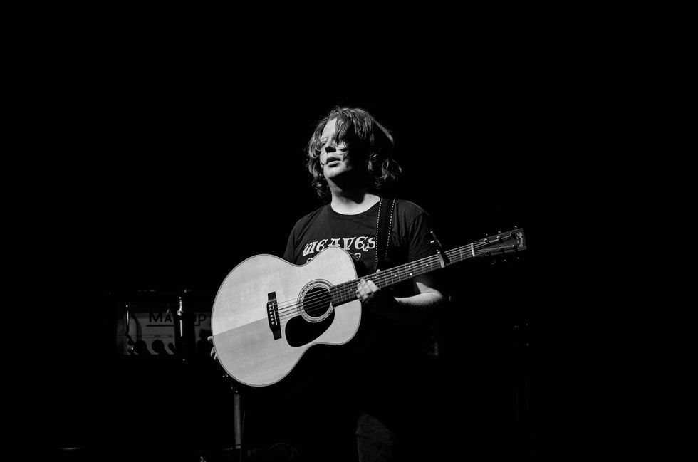 Musician with long hair holding an acoustic guitar on a dark stage.