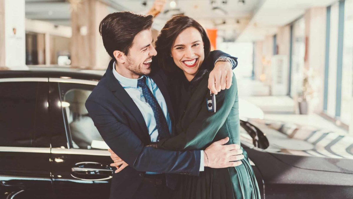 A young couple hug each other and smile at the camera, standing in front of their brand new luxury car.