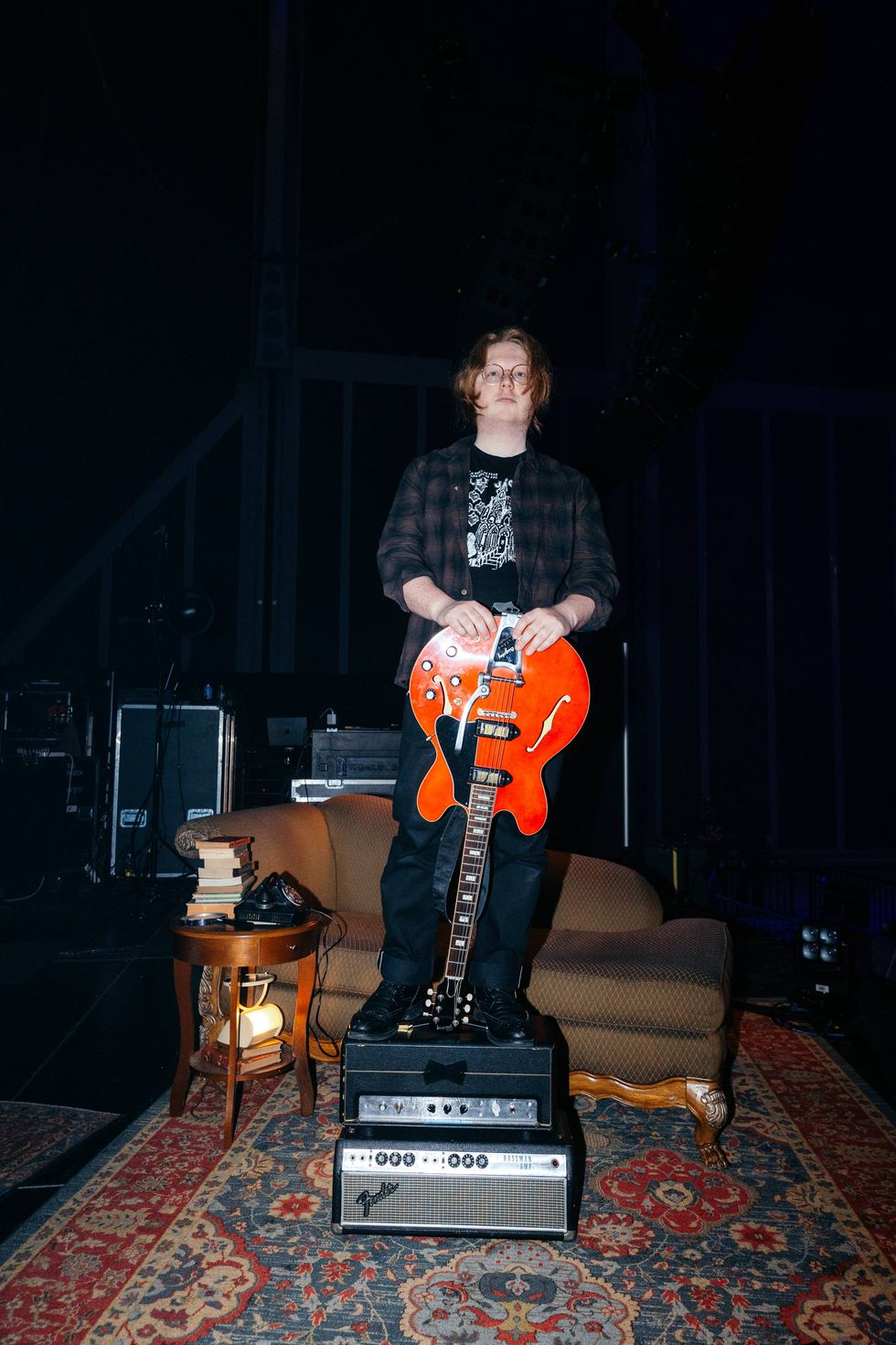 Person standing on amplifiers, holding a red guitar, in a dimly lit stage setting.