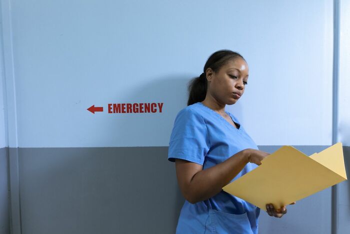 An ER staff member in scrubs reviewing files, standing next to an emergency sign, managing ridiculous patients.