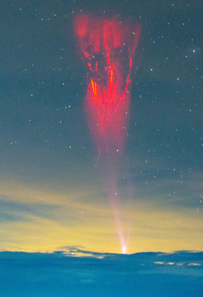 Lightning spreads like roots from a tree up into the atmosphere from the top of a cloud. Stars are scattered across the night sky above and the desert sits below the cloud deck.