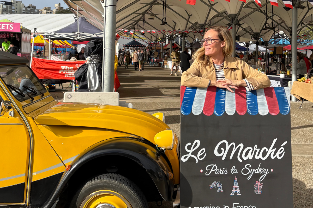 Woman stands next to yellow vintage car and a sign for the Le Marché markets.