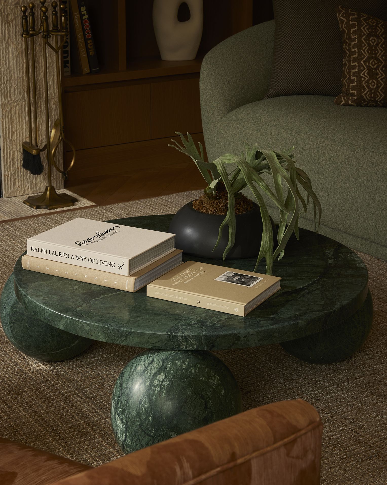 A green stone round coffee table with spherical legs, topped by three beige books and a black vase filled with a plant, placed next to green and brown armchairs.