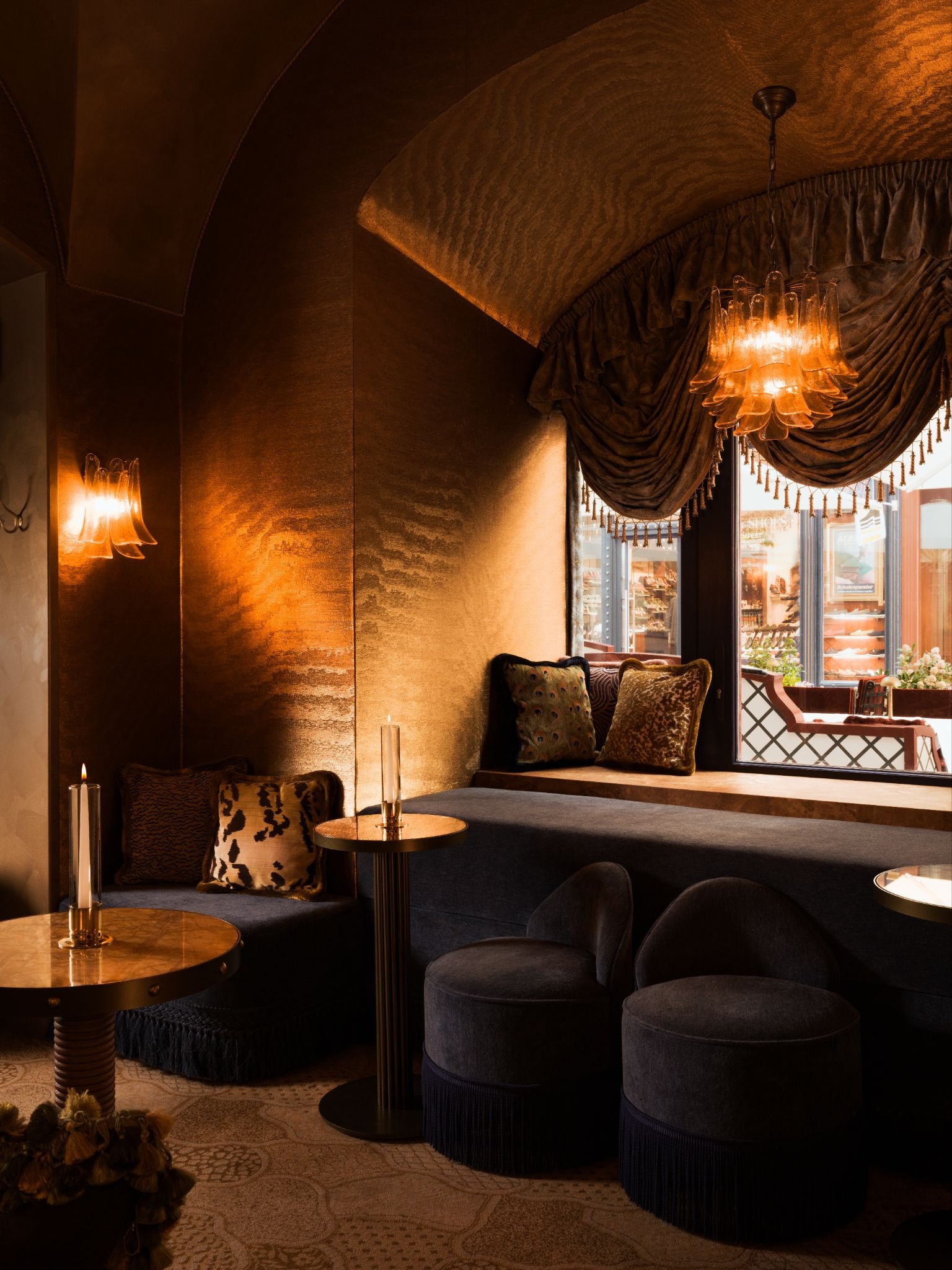 The interiors of a plush cocktail bair with velvety red, golden, and green rooms decorated with glossy chandeliers, golden sconces, opulent drapery, and intricate patterns.