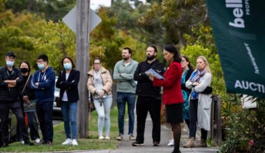 People standing on a footpath for a house auction. A woman in front of them is holding a folder.