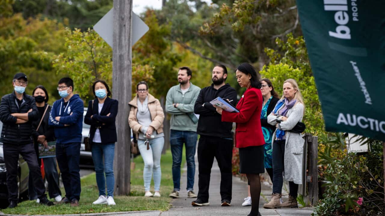 People standing on a footpath for a house auction. A woman in front of them is holding a folder.
