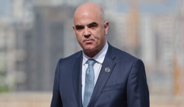 A medium close-up shot of Alain Berset, wearing a dark blue suit, a white shirt, a light blue patterned tie, and a small round lapel pin, looking forward with a serious expression against a blurred outdoor background.