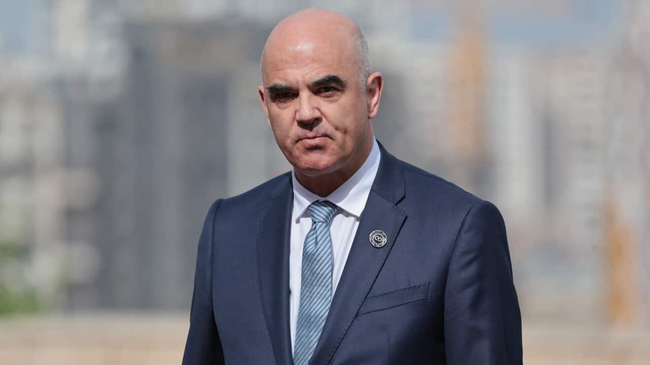 A medium close-up shot of Alain Berset, wearing a dark blue suit, a white shirt, a light blue patterned tie, and a small round lapel pin, looking forward with a serious expression against a blurred outdoor background.