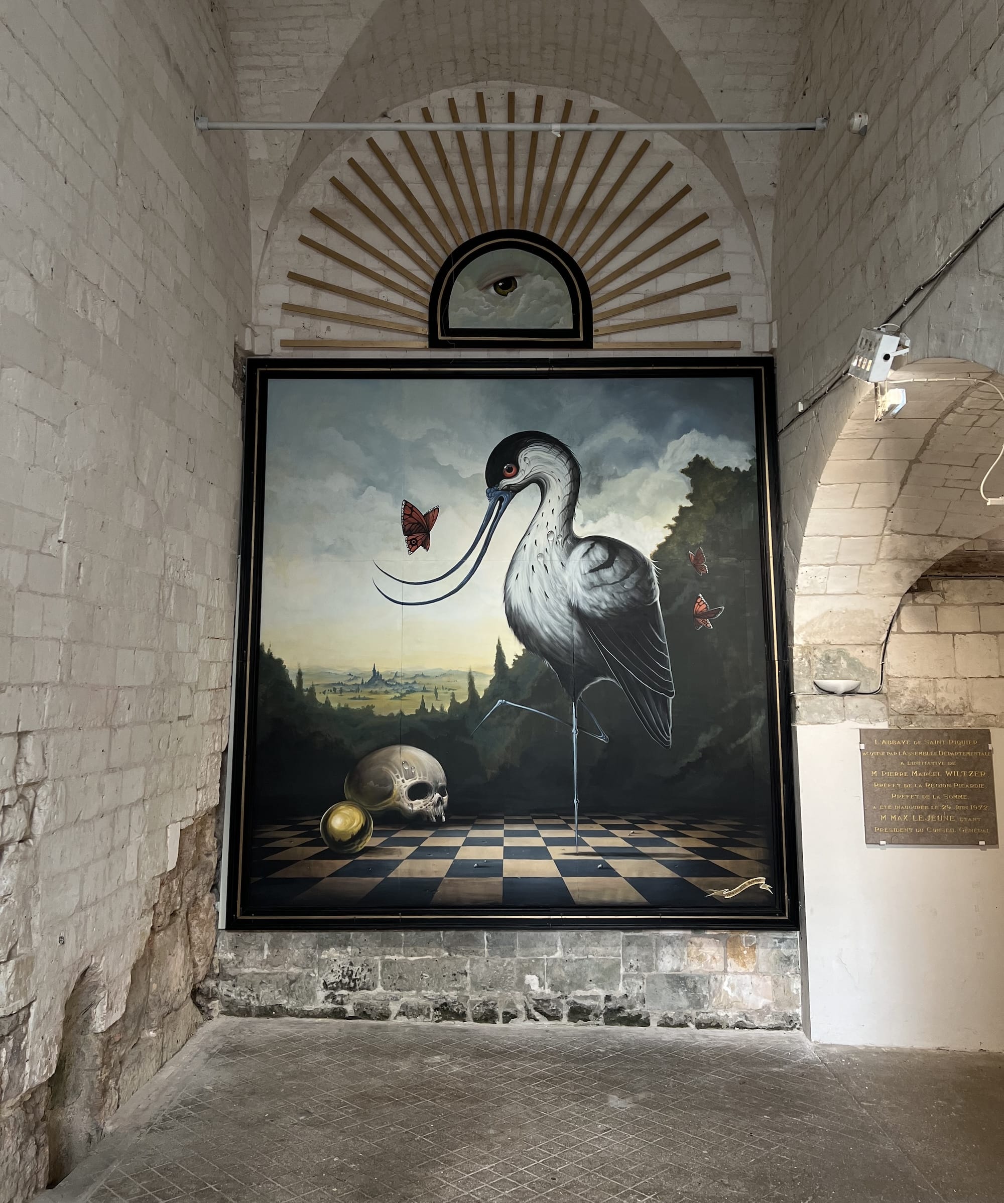 an interior mural of a bird, butterfly, and human skull on a checkered floor against a landscape, situated inside of historic church architecture