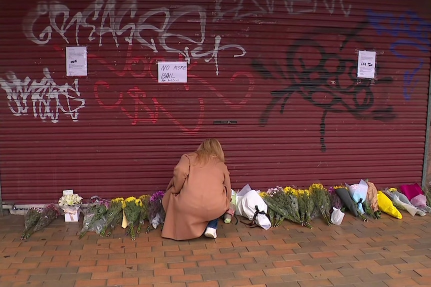 A woman in a camel coloured coat kneels on the footpath where flowers lay against a red roller door covered in graffiti.