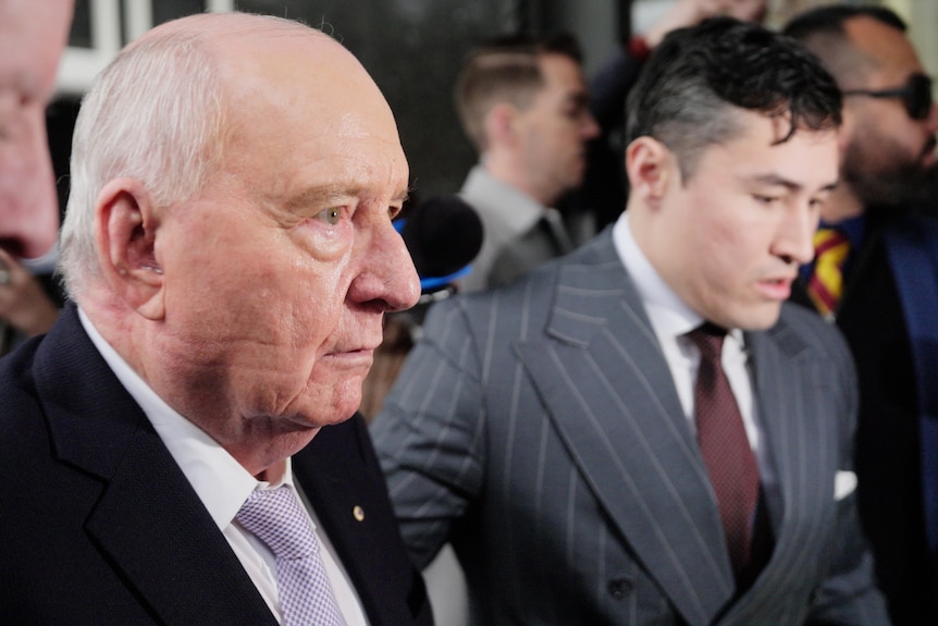 Alan Jones exits a car surrounded by men taking photos.