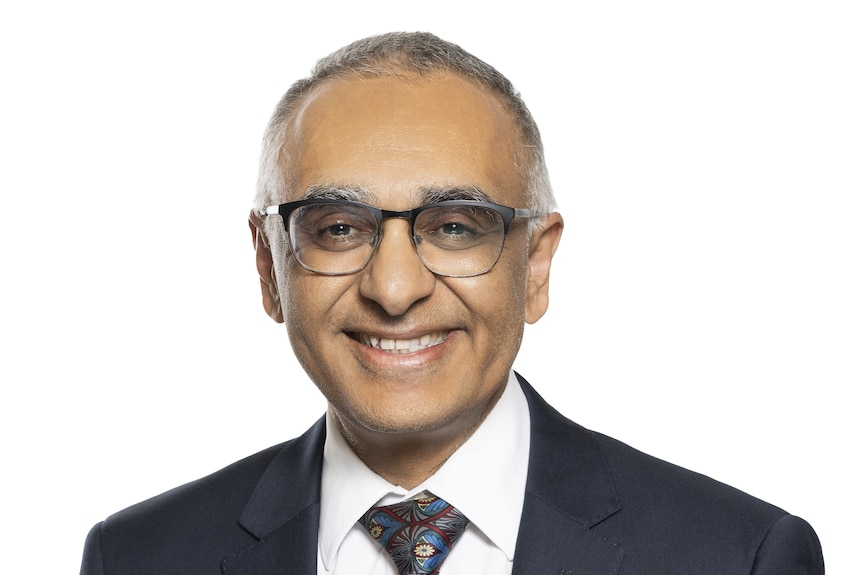 An older clean-shaven Indian gentleman wearing glasses smiles at the camera.