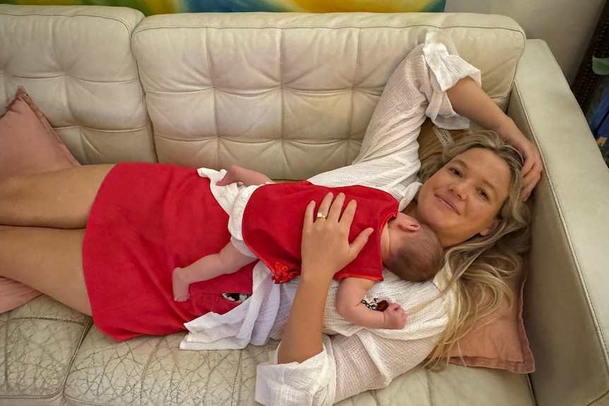 A blonde woman smiles, lying back with a small child in red on her chest.